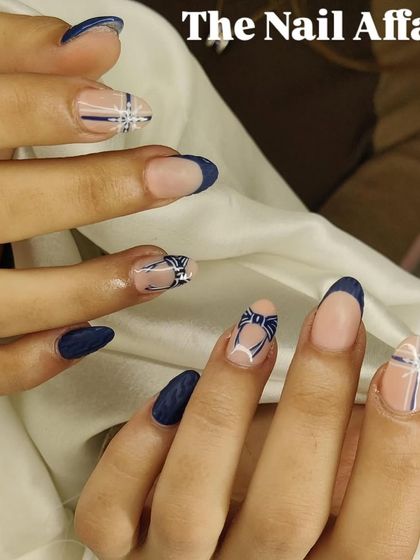 A unique and detailed design on almond nails, featuring a deep navy blue French tip combined with intricate, hand-painted bow and plaid patterns.