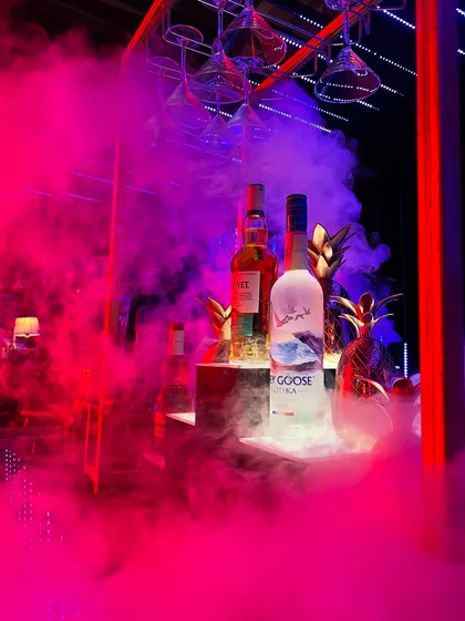 We use lighting and atmospheric effects to set the mood. Here, vibrant pink smoke envelops premium spirits like Grey Goose, creating a visually stunning and high-energy vibe for a modern cocktail party.