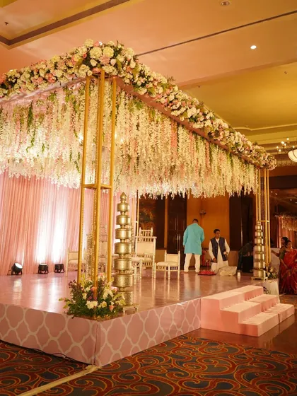 A classic mandap design with a canopy of cascading white and pink flowers, a soft pink draped background, and traditional golden pillars.