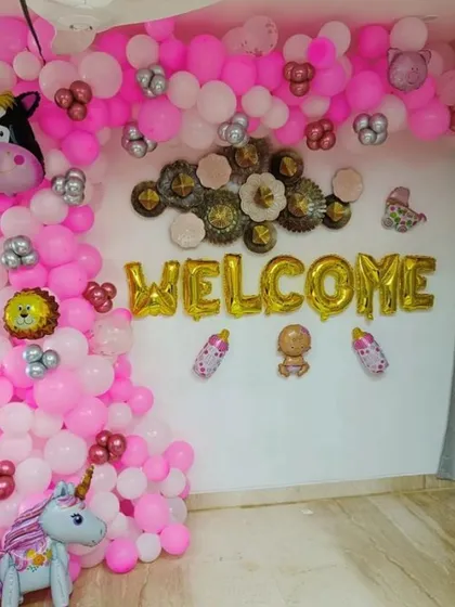 A 'Welcome' decoration for a baby girl. The wall is adorned with pink and white balloons, foil letters, and cute animal foil balloons like a unicorn and cow.