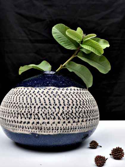 A finished, rounded pot with a dotted texture and a deep blue glaze. It serves as a perfect vase for a simple green sprig, bringing nature indoors.