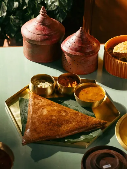 A crispy ghee roast dosa with sambar and chutney. The moody lighting and traditional props create a rich, atmospheric scene.