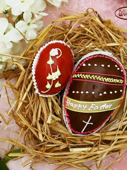 Two of our intricately decorated marzipan Easter eggs, nestled together in a straw nest. One is a vibrant red, while the other is a rich chocolate brown, both showing our detailed handiwork.