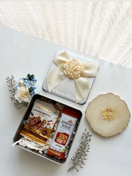A thoughtful return gift set including a tin of gourmet snacks and a beautiful resin coaster with gold leaf details.