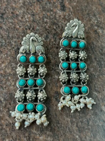 Pure silver earrings featuring a peacock motif and vibrant turquoise stones. The design is finished with delicate pearl drops, adding a touch of elegance and movement.