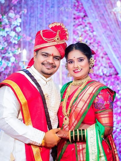 A classic portrait of the newlyweds on stage against a vibrant floral backdrop. The groom is wearing a traditional pheta (turban), and the bride is in a beautiful red and green saree, representing the complete Maharashtrian bridal look.
