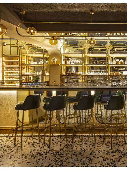 The main bar at Klap is a statement piece, with a brass structure, black leather stools, and intricate shelving. The design is both glamorous and functional, serving as the energetic heart of the restaurant.
