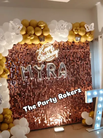 A beautiful 7th birthday setup with a rose gold sequin backdrop. The white and gold balloon arch is decorated with butterfly cutouts, and a large marquee number '7' adds to the sparkle.