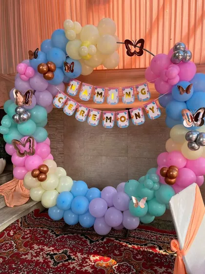 A pastel-colored Naming Ceremony decoration. This setup features a circular balloon arch decorated with butterfly cutouts and a "Naming Ceremony" banner across the center.