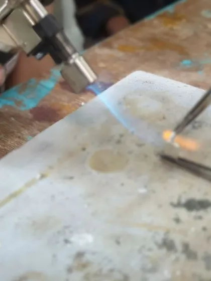 A close-up of a blowtorch heating a small piece of silver, demonstrating the soldering process used to join different parts of a jewelry piece together.