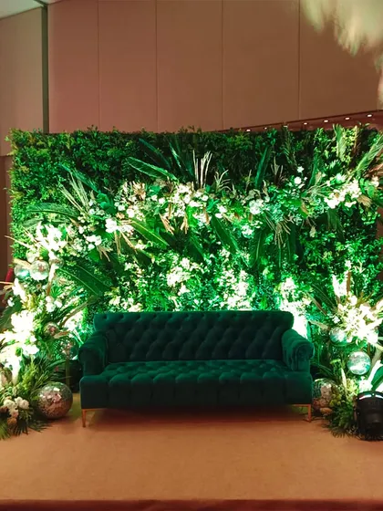 A vibrant green photo spot for a tropical or jungle-themed party. The emerald green velvet sofa stands out against a dense wall of foliage, lit to highlight the textures of the different leaves.