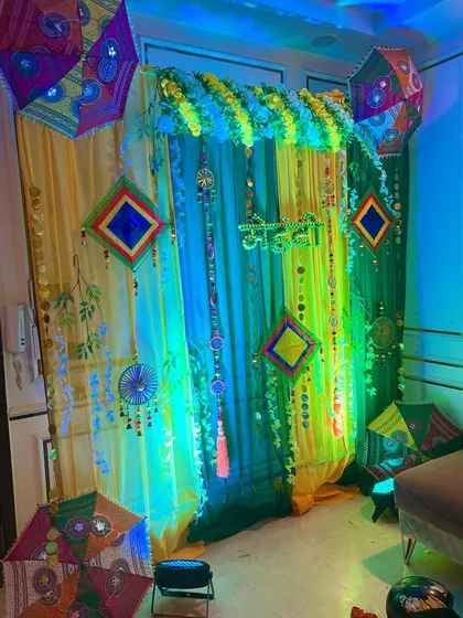 A Haldi or Mehndi backdrop with yellow and green drapes, marigold garlands, and ethnic umbrellas, illuminated with colorful lights.