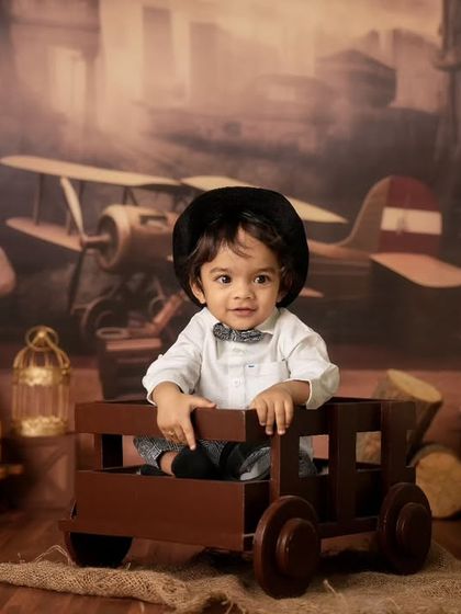 A toddler in a bow tie and hat sits in a wooden cart, posed against a vintage-themed backdrop featuring an old airplane.