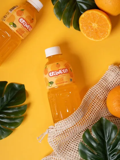 An orange-flavored drink styled with fresh oranges and monstera leaves in a mesh bag. This composition creates a fresh-from-the-market, natural, and summery vibe.