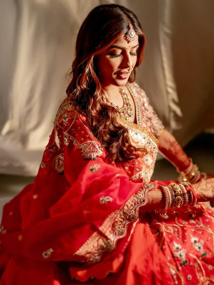 Bathed in warm sunlight, the bride's red lehenga and glowing makeup look even more radiant. This shot captures a beautiful, candid moment of her looking down.
