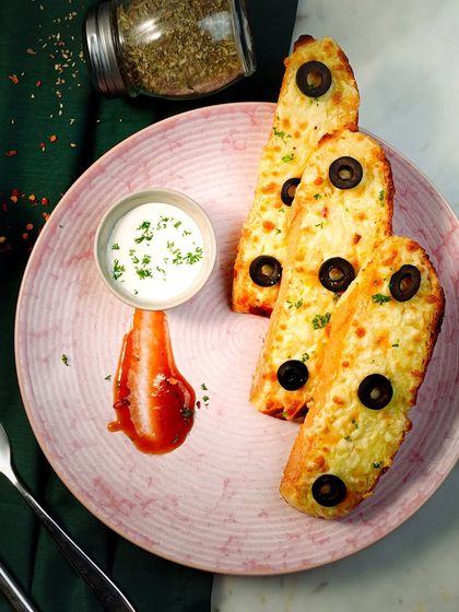 A beautifully plated shot of cheesy garlic bread with olives. The dark green background and elegant pink plate create a sophisticated, restaurant-quality look.