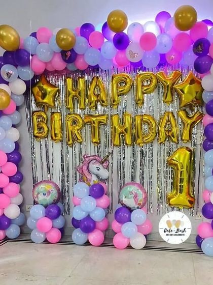 A unicorn-themed first birthday party with a purple, pink, and blue balloon arch against a silver fringe backdrop.