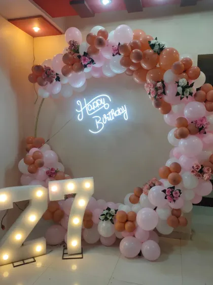 Celebrate a 27th birthday with our popular ring backdrop. This design features a circular frame adorned with pastel pink and rose gold balloons, faux flowers, a 'Happy Birthday' neon sign, and large light-up marquee numbers.