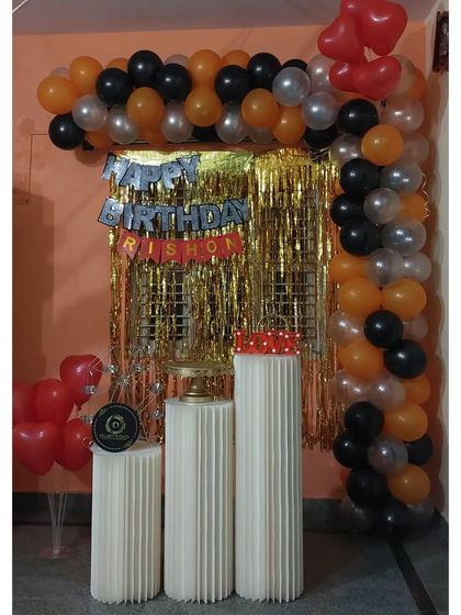 A simple and budget-friendly decor option for an at-home celebration. This setup uses an orange, black, and silver balloon garland with a gold tinsel curtain and pedestals for the cake.
