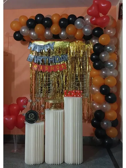 A simple and budget-friendly decor option for an at-home celebration. This setup uses an orange, black, and silver balloon garland with a gold tinsel curtain and pedestals for the cake.