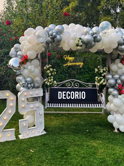 For a milestone 25th anniversary, I created this elegant outdoor swing decoration. The setup features a lush silver and white balloon arch, a custom 'Happy Anniversary' neon sign, and large marquee numbers, making it a beautiful focal point for photos in a garden or lawn setting.