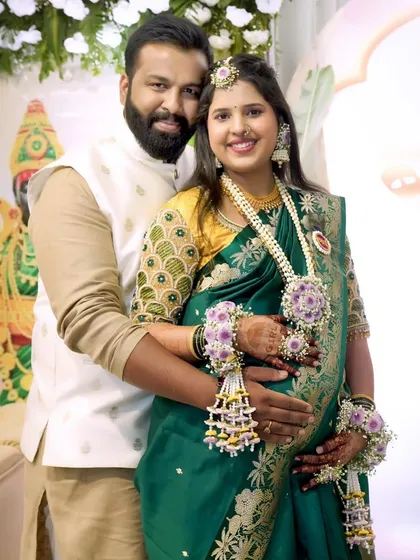 A happy couple at their baby shower. The mom-to-be is adorned in a custom set with purple flowers, featuring a maang tikka, necklace, and matching hand pieces.