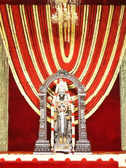 A stunning backdrop of red and gold floral garlands frames the sacred deity. Paired with a glowing chandelier, this design radiates divine energy and creates an atmosphere of pure elegance and grace.