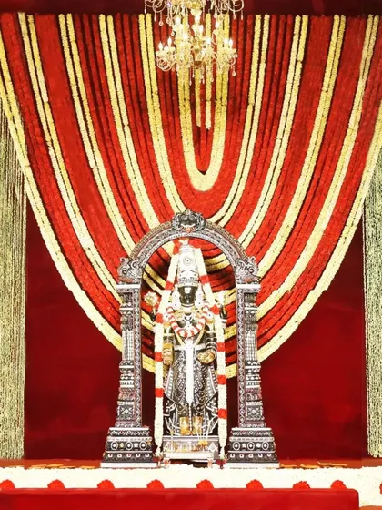 A stunning backdrop of red and gold floral garlands frames the sacred deity. Paired with a glowing chandelier, this design radiates divine energy and creates an atmosphere of pure elegance and grace.