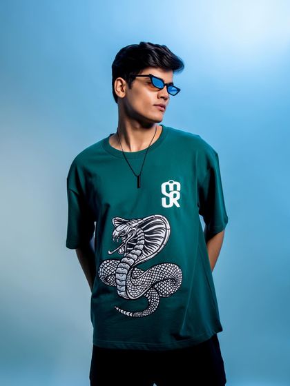 A vibrant shot with a blue background that makes the green graphic tee pop. The styling with slim sunglasses adds a trendy, modern touch to the overall look.