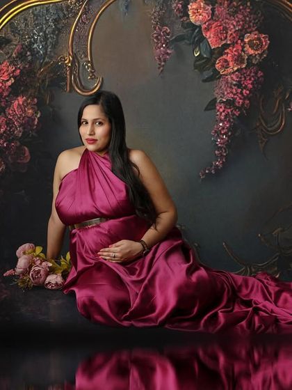 A moment of serene beauty. The mom-to-be is seated gracefully in a magenta gown against a painterly floral backdrop, her reflection adding to the artistry.