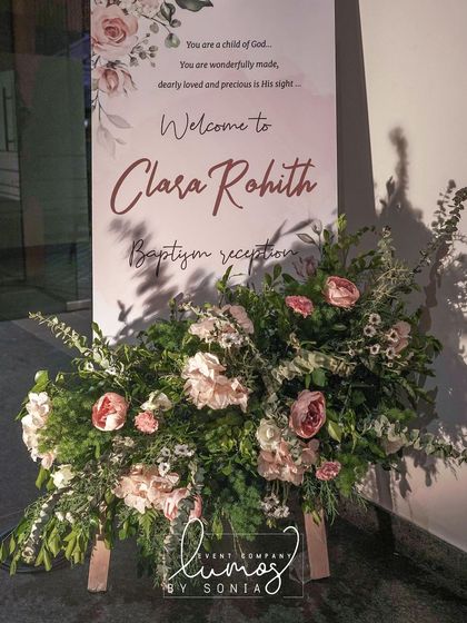 A close-up of the welcome sign for Clara Rohith's baptism reception, surrounded by lush pink and white florals.