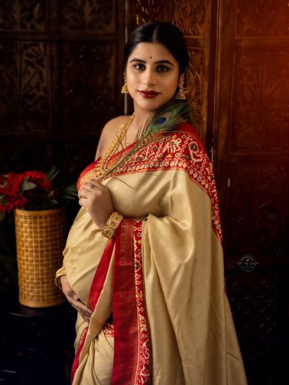 A graceful portrait of a mom-to-be in a cream and red silk saree, holding her bump with a peacock feather adding a touch of divine symbolism.
