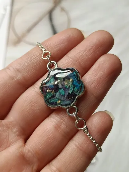 Holding a flower-shaped bracelet charm made from orchid petals. You can see the intricate details and colors of the flower preserved forever in the resin.