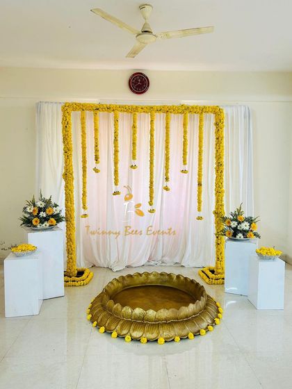 A simple and elegant Haldi backdrop with a marigold arch and hanging floral strings. This setup is ideal for creating a beautiful photo corner in any home space.