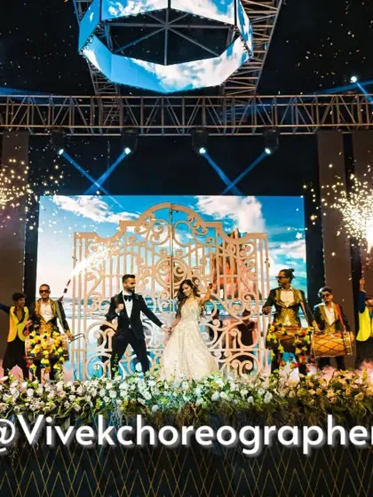 A wide shot of a spectacular sangeet entry. The couple stands center stage, framed by dancers with pyrotechnics and dhol players, creating a powerful and energetic visual.