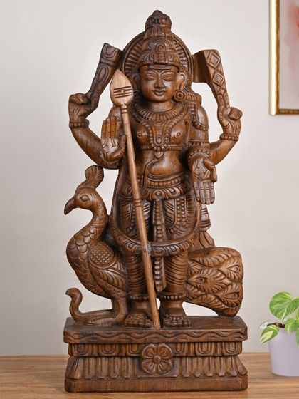 A 24-inch wooden Murugan idol with a classic wax brown finish. The intricate carving details his divine attributes, including his peacock vahana and the Vel.