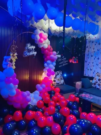 A vibrant room decoration with a mix of red, blue, pink, and black balloons covering the floor. A dreamcatcher and neon sign add to the eclectic and fun atmosphere.