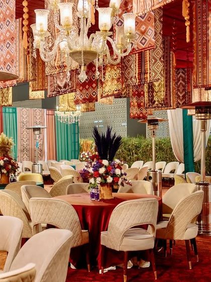 A luxurious vintage-themed event with a ceiling of hanging rugs, crystal chandeliers, and elegant guest seating in shades of cream, red, and teal.