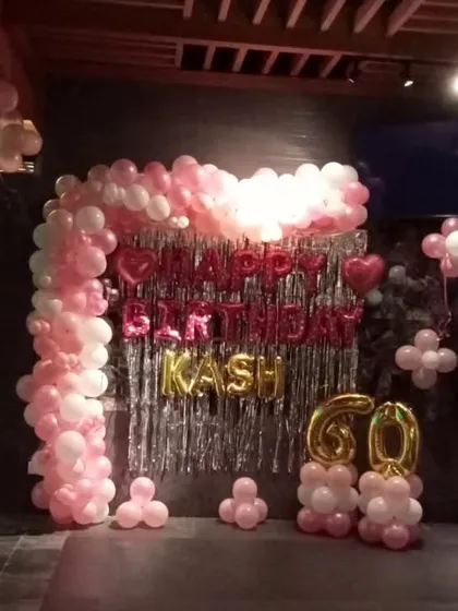 A 60th birthday party at a restaurant, decorated with a pink and white balloon arch over a silver fringe backdrop and large foil number balloons.