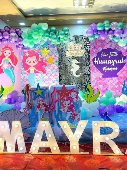 A vibrant 'Our Little Mermaid' party with large marquee letters spelling out the name 'Humayra'. The stage features character backdrops, a shimmer wall, and a colorful balloon arrangement.