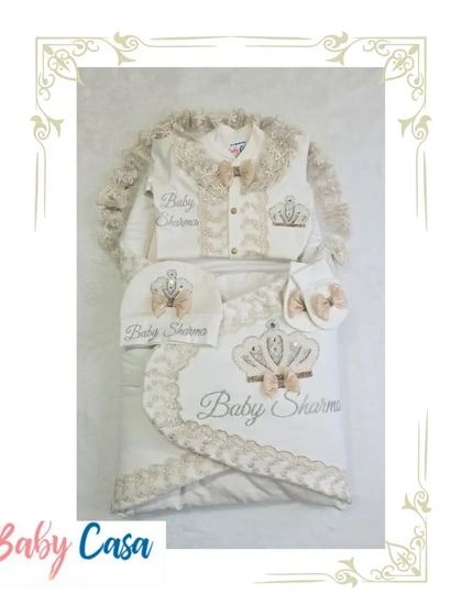 A complete personalized set for "Baby Sharma." This neutral toned collection includes a wrap, romper, cap, and bib, all featuring a beautiful crown motif with a bow.