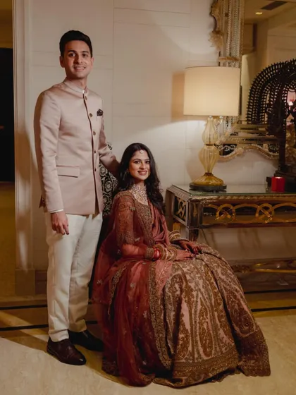 A formal portrait of the couple. The bride is seated, showcasing the beautiful paisley print on her lehenga, while the groom stands beside her in his perfectly fitted bandhgala, a picture of classic elegance.