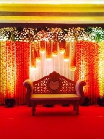 This stage design uses hanging flower strings and warm lighting to create a glowing backdrop. The traditional wooden sofa completes this setup, ideal for a sangeet or wedding reception.