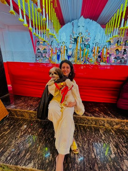 A pet parent holds her adorable Shih Tzu, both posing happily in front of the decorated Durga idol. It's a beautiful moment of shared celebration and blessings.