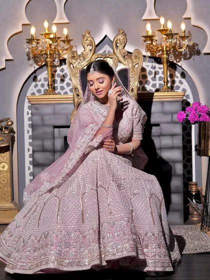A majestic shot of the bride seated on a throne-like chair. The full view of her lilac-pink lehenga and the grand setting is complemented by her flawless, regal makeup.