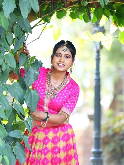 A smiling shot of the model in the pink lehenga, peeking through green leaves, showcasing a fun and festive vibe.