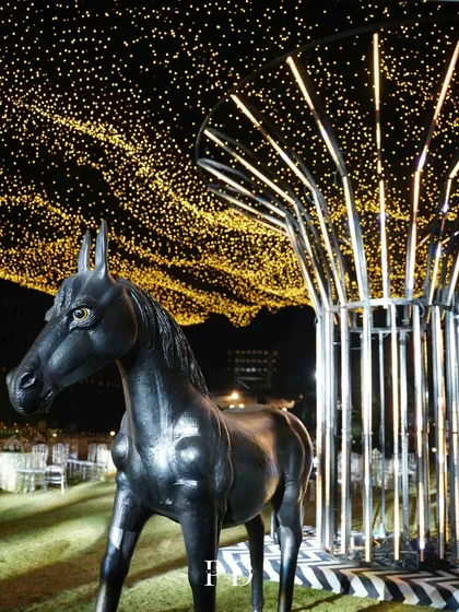 A unique and artistic decor element for a Sangeet, featuring a black horse statue under a modern light installation and a canopy of fairy lights.
