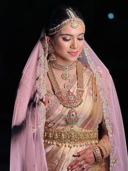A stunning bride in a golden saree with a sheer pink veil. The makeup features a soft pink eyeshadow and a matching lip, creating a dreamy, romantic look.