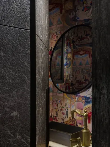 A peek into the powder room of the Caitriona apartment, revealed through a creatively designed stone door. The room features playful 'Cole & Sons' wallpaper depicting cabaret dancers, adding a touch of whimsy and flair.