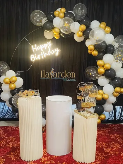 A stylish half-ring balloon arch in black, white, and gold. This setup includes a neon sign and multiple pedestals for cake and desserts, perfect for a chic celebration.